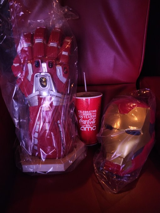 An Iron Man themed movie theater promotional setup featuring a large gauntlet replica, a red and gold helmet, and a Coca-Cola AMC cup celebrating 100 years.
