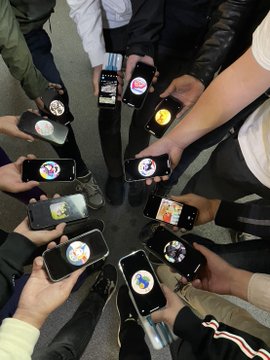 The image shows a group of people holding up their cell phones, with each person displaying their phone's screen. The scene appears to be set at an event or gathering where attendees are showcasing their devices. The focus of the image is on the various smartphones held by the individuals in the circle, highlighting the modern technology and its impact on social interactions and communication.