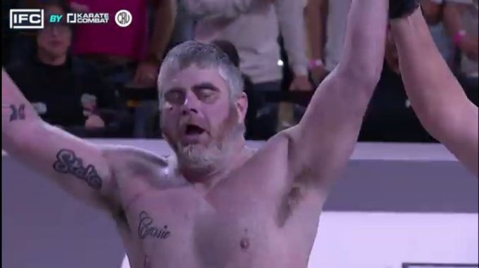A shirtless, bearded male fighter with tattoos (including 'State' and 'Caution') raising his arms in victory at a combat sports event with 'IFC' and 'Karate Combat' logos visible.