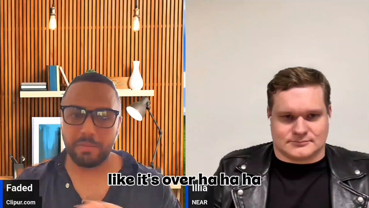 The image shows two men sitting next to each other with their faces displayed on a computer screen during an interview. They are both wearing glasses, and there's a microphone attached to the camera capturing their conversation. The background of the image features wooden panels, which adds a rustic touch to the scene. The focus of the image is on the two men engaged in a discussion or interview, with one man appearing more serious while the other seems to be making a joke.