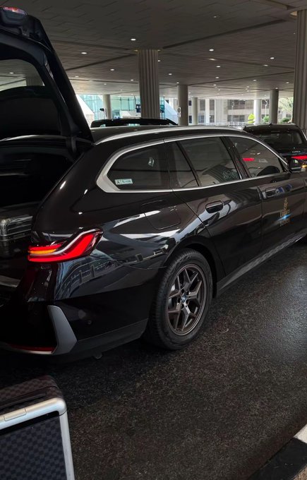 A sleek black BMW station wagon is parked at a hotel entrance with its trunk open, displaying its polished exterior and chrome trim. The image juxtaposes the modest, real-world luxury of an everyday vehicle with the post's caption about "generational wealth" from a memecoin, highlighting the aspirational contrast between crypto dreams and tangible possessions.