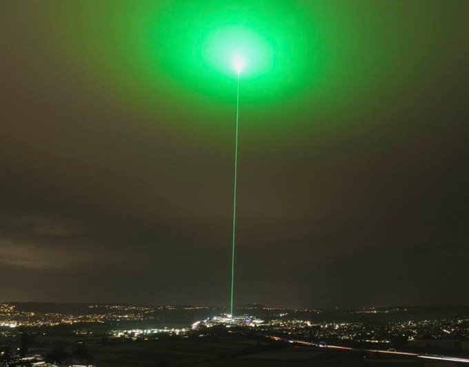 A powerful green laser beam shoots skyward from a city at night, forming a bright glowing orb overhead like a giant bullseye — perfectly mirroring the "crypto god candles are coming" post as a visual metaphor for a massive bullish signal piercing the market.