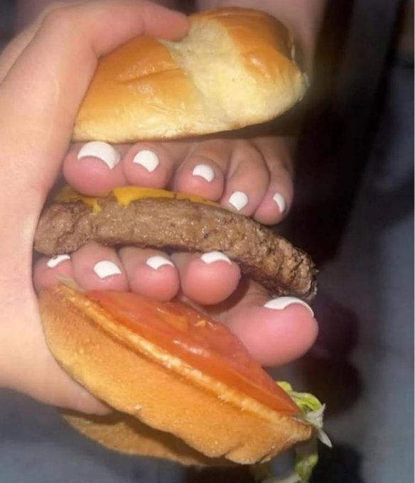 A hand grips a cheeseburger so tightly that the toes—with white-painted toenails—press out from between the bun, patty, and tomato, making it look as though the foot is "eating" the burger in a visual pun on "gtoes."