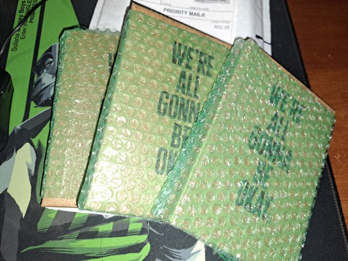 Three packages wrapped in green bubble wrap with text including 'WE'RE ALL GONNA BE OK' and 'YOU'VE GOT THIS', sitting on a priority mail envelope.