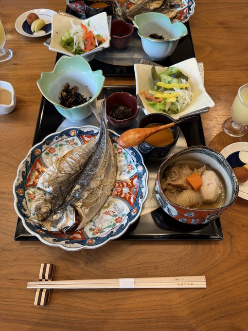A traditional Japanese meal set on a wooden table including a whole grilled fish on a decorative plate, miso soup, salads, pickles, and chopsticks on a rest.