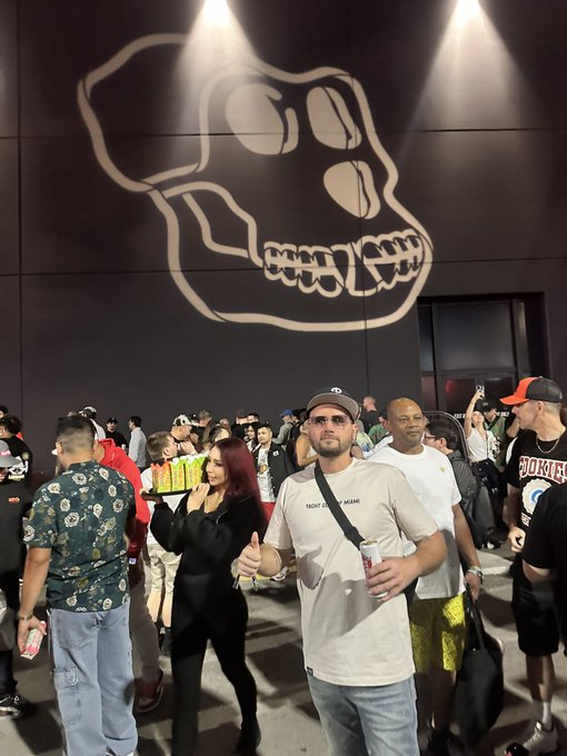 A group of people gathered at an event in front of a large skull graphic on the wall, including a man in a white 'Talent Don't Dream' t-shirt holding a can.