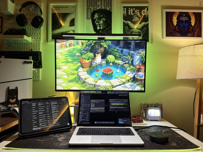 A desk setup featuring a monitor displaying a garden scene, a laptop showing code, a tablet, headphones, and posters including 'Doctor' and 'it is d' on the wall.