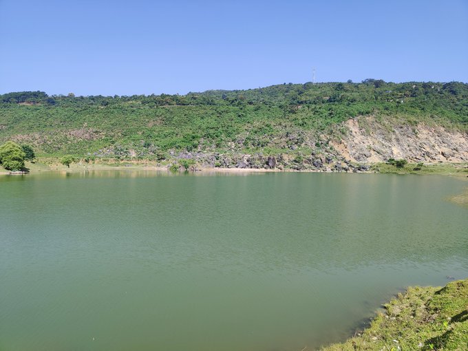 A serene lake with greenish water sits at the base of a lush, tree-covered hillside under a clear blue sky, with what appears to be a communications tower visible on the distant ridge. The image's calm natural landscape contrasts with the post's message, possibly symbolizing the peaceful, low-impact energy efficiency that the quantum network project aims to achieve compared to traditional power-hungry mining operations.