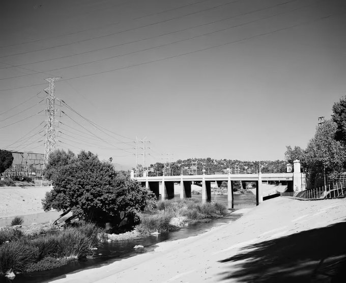 A black and white photograph of a concrete bridge spanning a dry concrete riverbed, with power lines and transmission towers in the background under a clear sky.