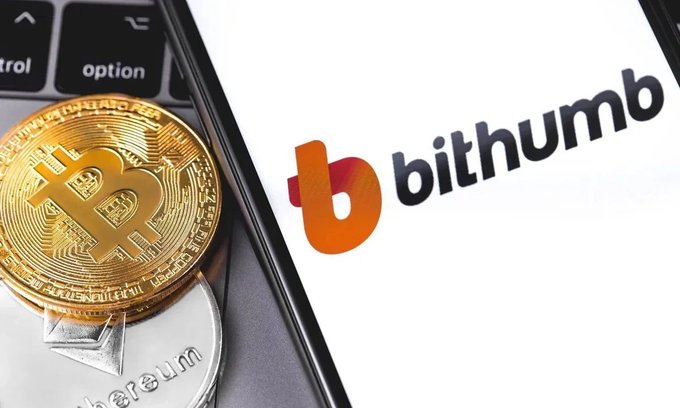 A close-up photo of a physical Bitcoin token resting on a laptop keyboard next to a smartphone displaying the Bithumb cryptocurrency exchange logo.