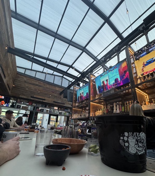 An interior view of a sports bar with a glass roof, multiple TVs displaying content, and a bar counter with drinks, a bucket, and bowls in the foreground.