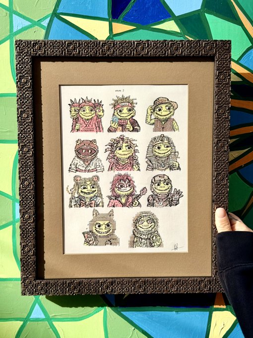 An ornately framed cross-stitch artwork titled 'Vibe 1' depicting 10 whimsical character portraits, held up against a colorful stained-glass background.