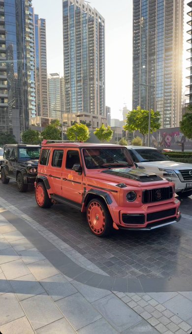 A bold, custom matte-red Mercedes-Benz G-Wagon—featuring a blacked-out hood and matching red alloy wheels—is parked prominently in an urban setting framed by towering glass skyscrapers, with a black G-Wagon beside it and a white SUV to its rear. The post's tongue-in-cheek caption, "GM to everyone who says GM," plays on the vehicle's branding to deliver a stylish, confident greeting rather than referring to General Motors.