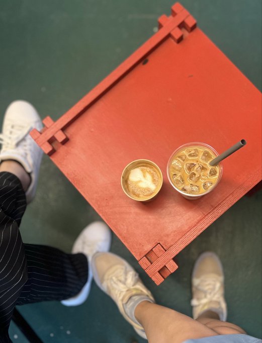 An overhead photo of two iced coffee drinks on a small red table, with two people wearing white sneakers and one in pinstriped pants standing around it.