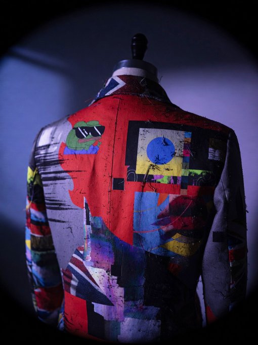 A dark, dramatic photograph shows the back of a mannequin wearing a denim jacket painted with colorful, abstract art, including a Union Jack on the collar and a cartoon frog wearing sunglasses.
