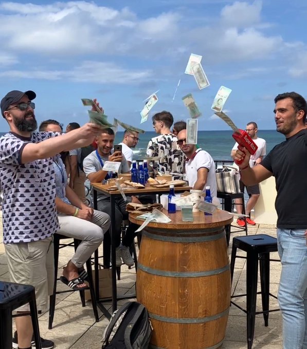 A group of men at a beachside gathering throw US dollar bills into the air, with one man on the right holding a red spray can.
