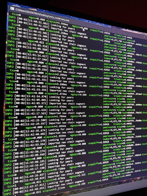 A computer monitor displays a command prompt window running mgas=0.000, a node.exe process on the C:\Users\SEEKTRU\Desktop directory, showing repetitive log entries with timestamps from 08-02 indicating peer connections and chain segment imports. The screen supports the claim of mining VeryChat Token, as the visible node activity aligns with cryptocurrency mining operations rather than malicious hacking.