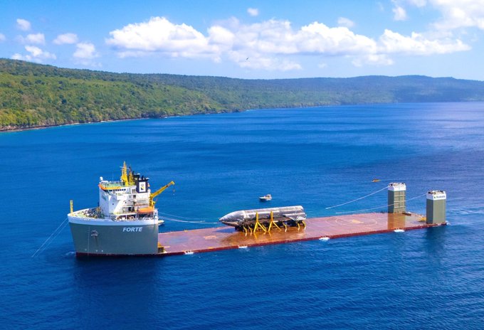 An aerial view of a large cargo ship named 'FORTE' transporting what appears to be a submarine on a red deck through blue ocean waters.