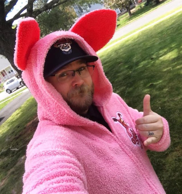 A bearded man wearing glasses, a police-style cap, and a pink fuzzy Piglet-style onesie with red ears gives a thumbs-up outdoors.