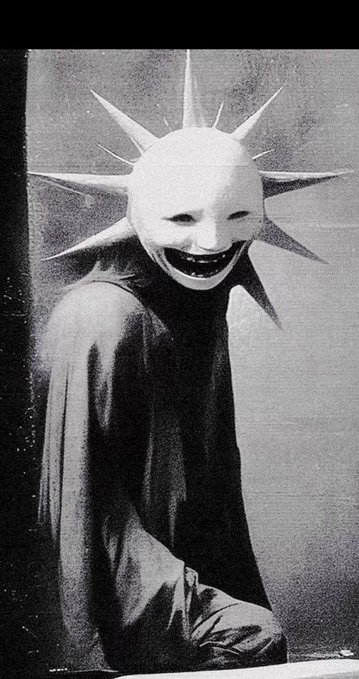 A grainy black-and-white image shows a sinister figure with a spiked, smiling mask draped in a dark cloak.