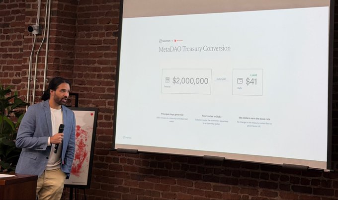 A man in a blue blazer presents a slide titled 'MetaDAO Treasury Conversion' showing $2,000,000 treasury converting to 1 Unit at $41 each, displayed on a brick wall.