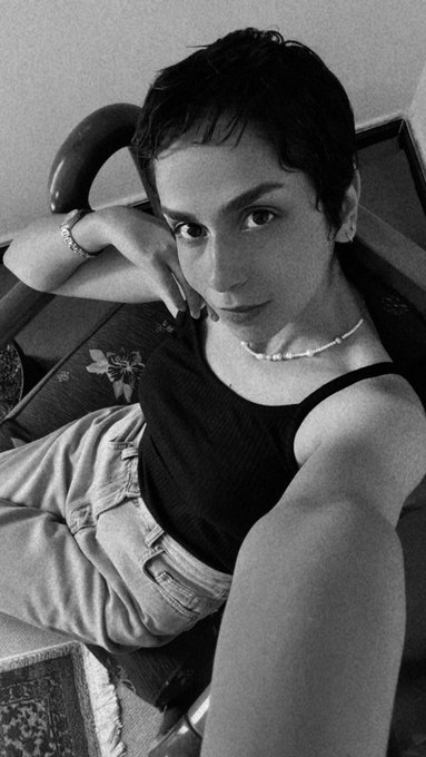 A black and white selfie of a young woman with short hair, wearing a dark tank top and a delicate pearl necklace, posing against a chair.