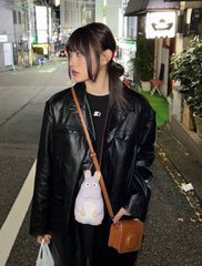A young woman with long dark hair wearing a black leather jacket and carrying a brown bag, standing on a city street.