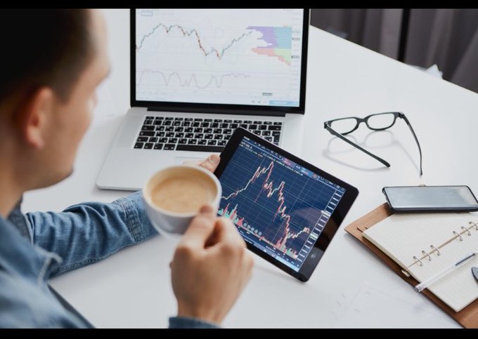 An over-the-shoulder view of a person holding a cup of coffee while analyzing stock market charts on a laptop and tablet on a white desk, with glasses and a notebook nearby.