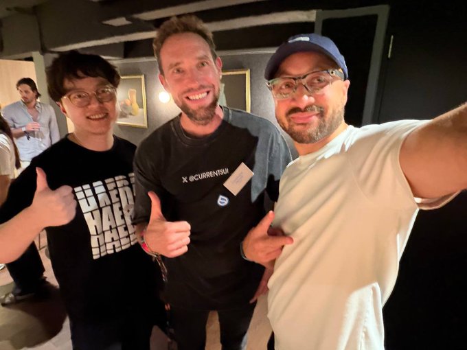Three men pose together for a selfie-style photo at what appears to be an event, with one wearing a shirt that reads 'X @CURRENTSUI'.