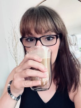 A selfie of a woman with brown hair and glasses drinking an iced coffee, with a small tattoo visible on her arm.