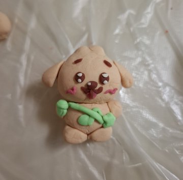 A small handmade clay figurine of a beige puppy with pink cheeks holding a green bone-shaped object, sitting on a plastic-covered surface.