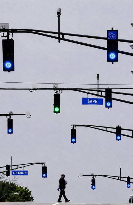 A man walks past numerous blue traffic signals and a green traffic light, with street signs labeled "$APE" and "APECHAIN" suspended overhead, playfully linking the visual flow of signals to the optimism in the caption "$APE."