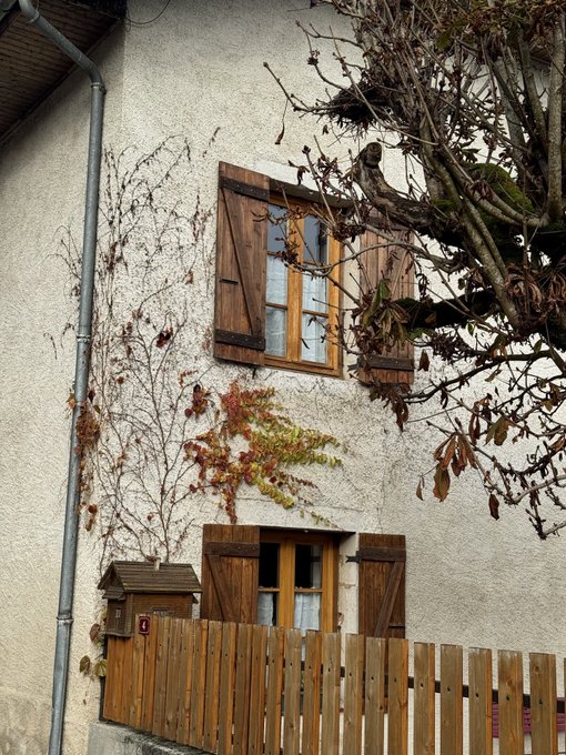 A rustic stone building with weathered wooden shutters and climbing vines stands behind a wooden picket fence, embodying the post's theme of builders keeping building amid market turbulence. The autumnal foliage and sturdy architecture serve as a visual metaphor for resilience and grounded progress when markets and narratives feel unstable.