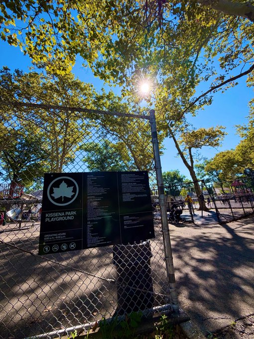 A sign reading 'KISSENA PARK PLAYGROUND' and 'DEPARTMENT OF PARKS AND RECREATION - QUEENS, NY' is attached to a chain-link fence with a playground and trees visible behind it.