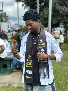 A young man wearing a 'CLASS OF 2024' sash stands outdoors holding several pens, surrounded by other people in what appears to be a gathering or celebration event.