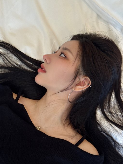 A young woman with long black hair lies on a bed, gazing upward, wearing a black top and delicate gold jewelry, creating a soft, contemplative aesthetic that complements the minimalistic Korean beauty tone of the USPEER/DAON post.