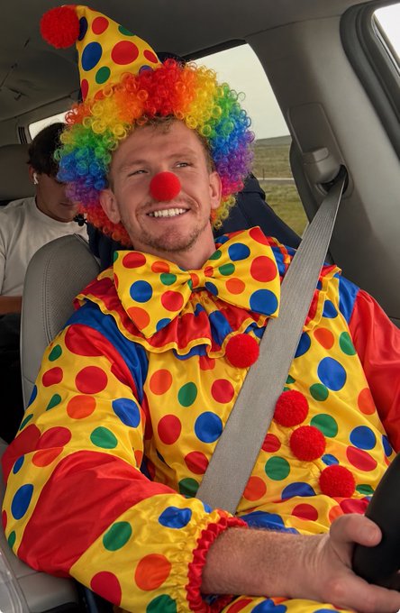 A smiling man dressed as a clown with a rainbow wig, red nose, and colorful polka-dot costume sits in the driver's seat of a car wearing a seatbelt.