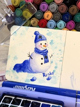 The image features a snowman made out of paper, sitting next to a collection of paint markers. The snowman has a blue scarf wrapped around its neck and a blue hat on top of its head. The markers are scattered around the snowman's base, with some in shades of purple and green. The post includes text that reads "Drew a little Rialo snowman today Just a cozy reminder that even when the grind feels like winter, this community’s always here keeping things warm".