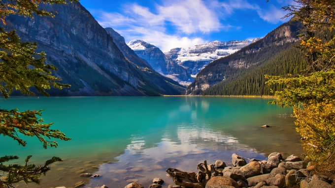 A stunning turquoise alpine lake (Lake Louise in Banff National Park, Canada) is framed by snow-capped mountains, evergreen forests, and autumn foliage, with rocky shoreline in the foreground. The image humorously reimagines the famous landmark as "Lake Satoshi," playing on Bitcoin's mysterious creator Satoshi Nakamoto and crypto enthusiasts' love of "moon" and lake metaphors.