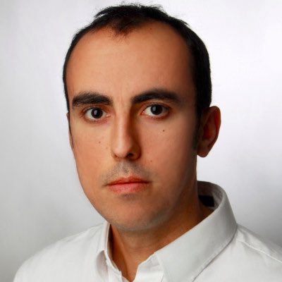 A close-up portrait photograph of a man with short dark hair, prominent eyebrows, and a neutral expression, wearing a white collared shirt against a plain light background.