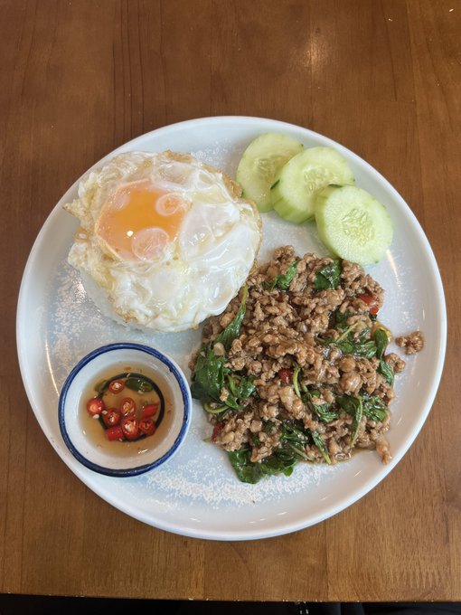 A white plate on a wooden table contains a fried egg with a runny yolk, sliced cucumbers, minced meat stir-fried with basil, and a small dish of fish sauce with sliced chilies.