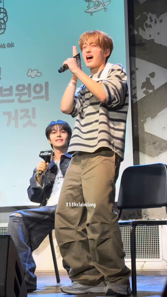 The image features two young men standing next to each other, with one of them holding a microphone while the other has his hands raised. They are both dressed in brown pants and white shirts. The background includes a blue wall with some writing on it, which appears to be related to the content of the post that says "2026/06/21 스테이지 ㄱㅇㅇ #ALPHADRIVEONE #ALD1 #알파드라이브원 ㄱㅇㅇ #씬롱 #XINLONG #シンロン #鑫隆 #상현 #SANGHYUN". The image seems to be related to the post, as it features two young men dressed in brown pants and white shirts.