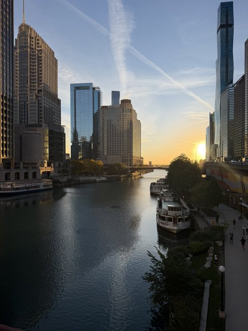A sunset view of the Chicago River flowing through downtown skyscrapers, with boats docked along the riverwalk and the sun setting on the horizon.