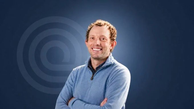 The image features a man wearing a blue sweater with crossed arms and a smile on his face. The background of the photo has a dark blue color that contrasts with the man's attire. There are no texts or other objects present in the image, making it a simple yet striking composition.