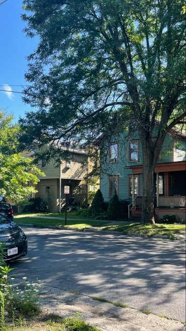 A photograph of a quiet residential street with large trees, featuring a two-story blue house and a parked car on a sunny day.