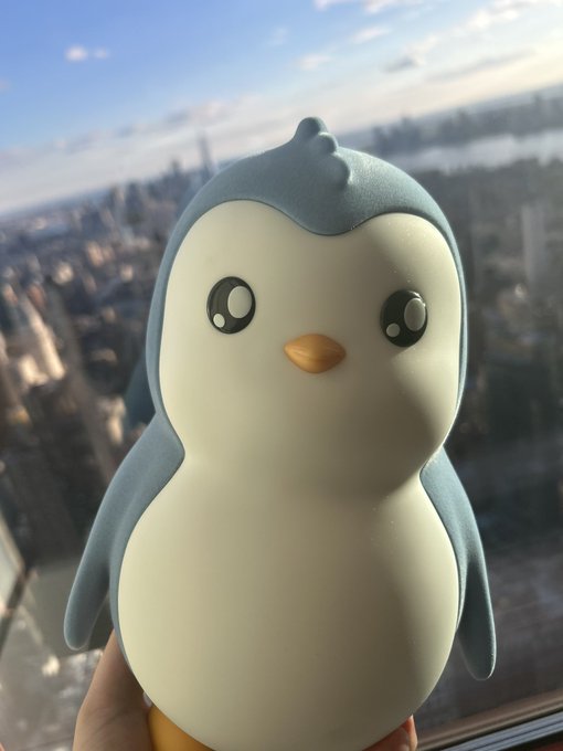 A Squishmallow-style plush penguin with big shiny eyes is held up against a window overlooking the New York City skyline, with the Hudson River visible in the background. The image captures the "Pengu" mascot atop a high-rise "Tokenization Tower," playfully tying the adorable penguin figurine to the soaring urban heights behind it.