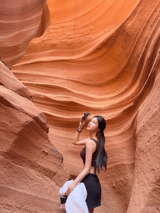A woman with long dark hair, wearing a brown crop top and black skirt, stands in a narrow slot canyon with undulating orange-red sandstone walls, holding a camera up to her face.