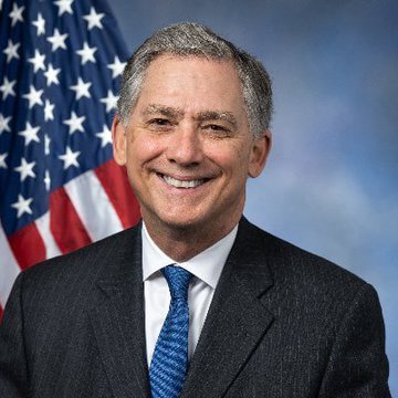 An official portrait of a smiling man in a dark suit and blue tie standing in front of an American flag.