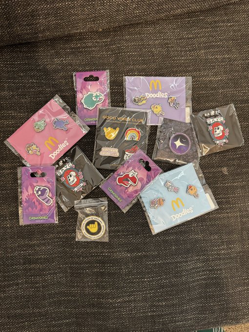 A collection of McDonald's-themed enamel pins in various packages featuring designs from collaborations with brands like Doodles, Bored Ape, and CleverMonkeys scattered on a dark fabric surface.