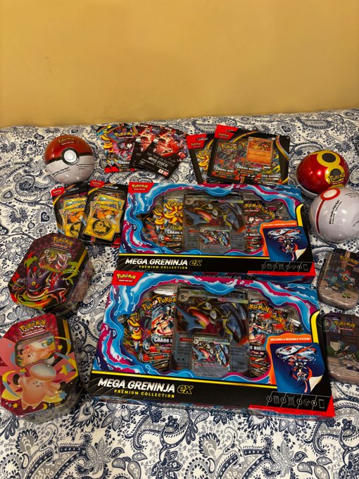 A Pokémon TCG collection spread out on a patterned bedsheet, including Mega Greninja ex Premium Collection boxes, booster packs, and various Poké Balls (standard, Great, Ultra, and Master Ball).
